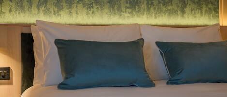 Habitación de hotel con cama, almohadas, un cuadro enmarco de un paisaje y una mesita de noche.