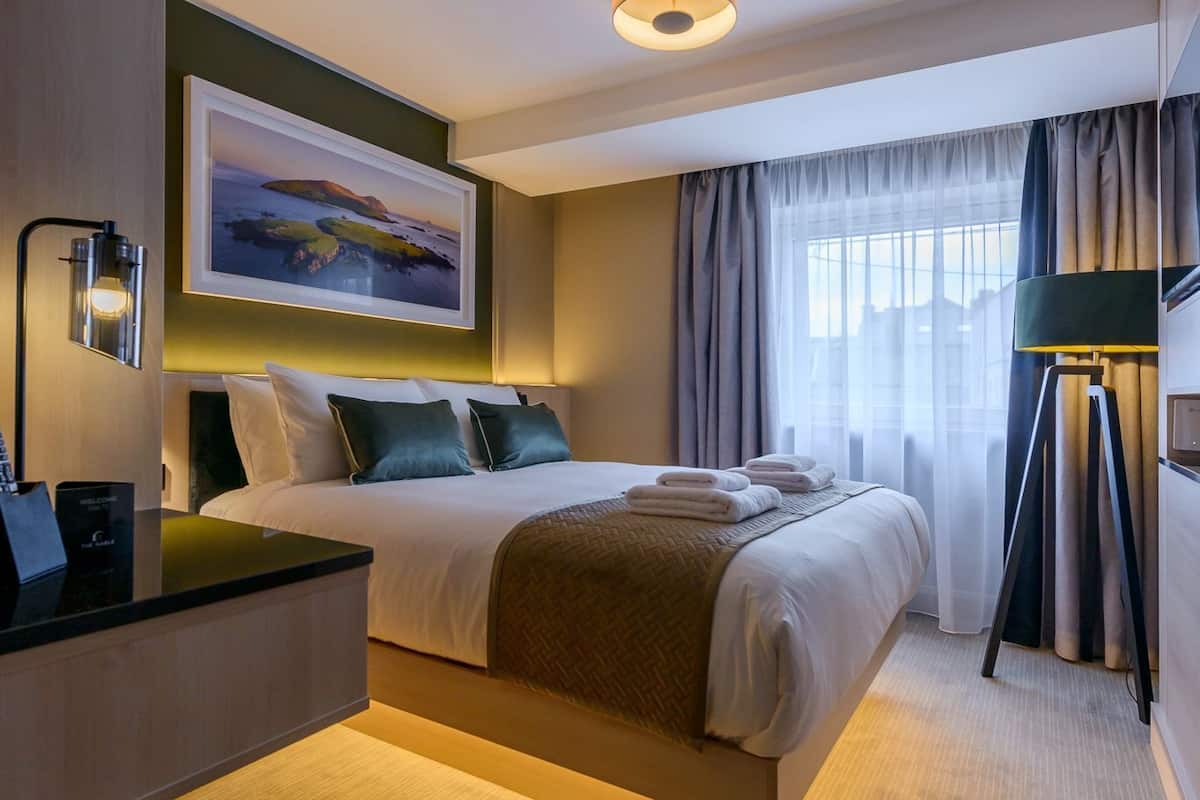 A hotel room with a bed, bedside table, lamp, and a painting on the wall.