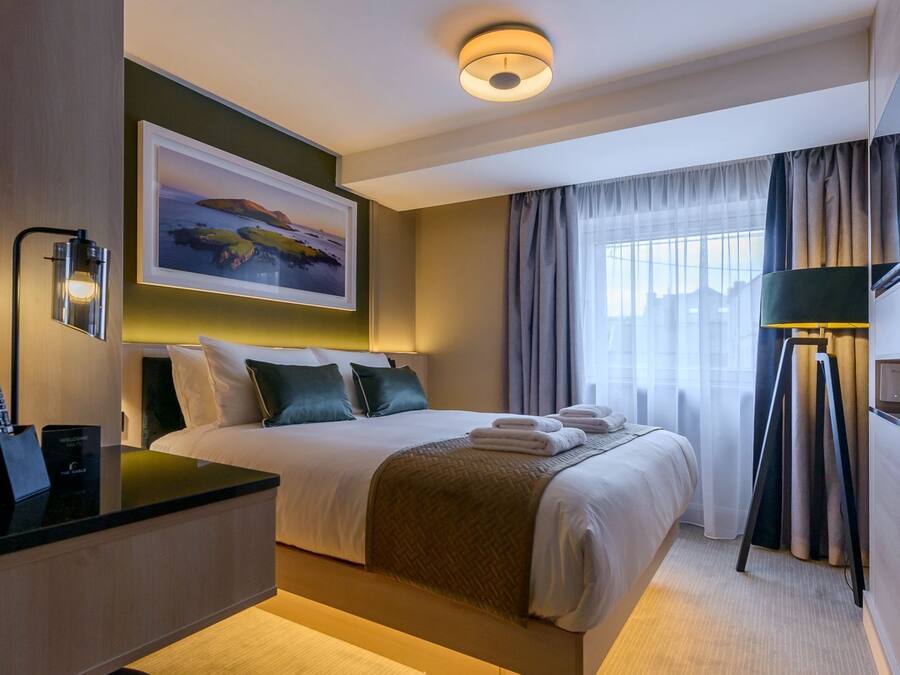 Una habitación de hotel con cama, mesita de noche, lámpara y un cuadro en la pared.