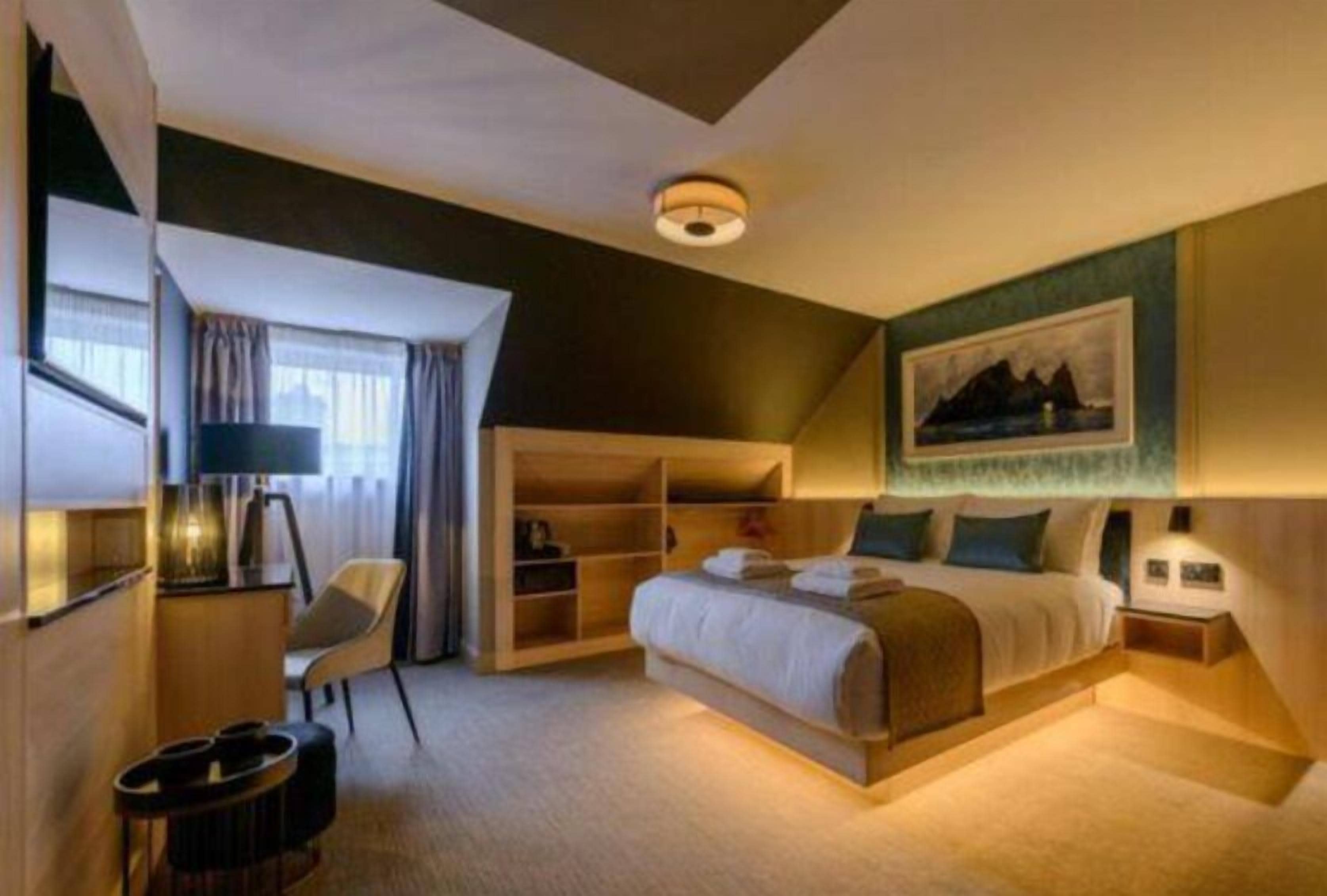 A hotel room with a large bed, a desk, a chair, a bookshelf, and a painting on the wall.