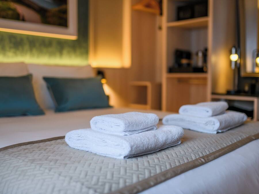 A neatly made bed with folded white towels, a woven bed skirt, and a bedside table with a lamp.