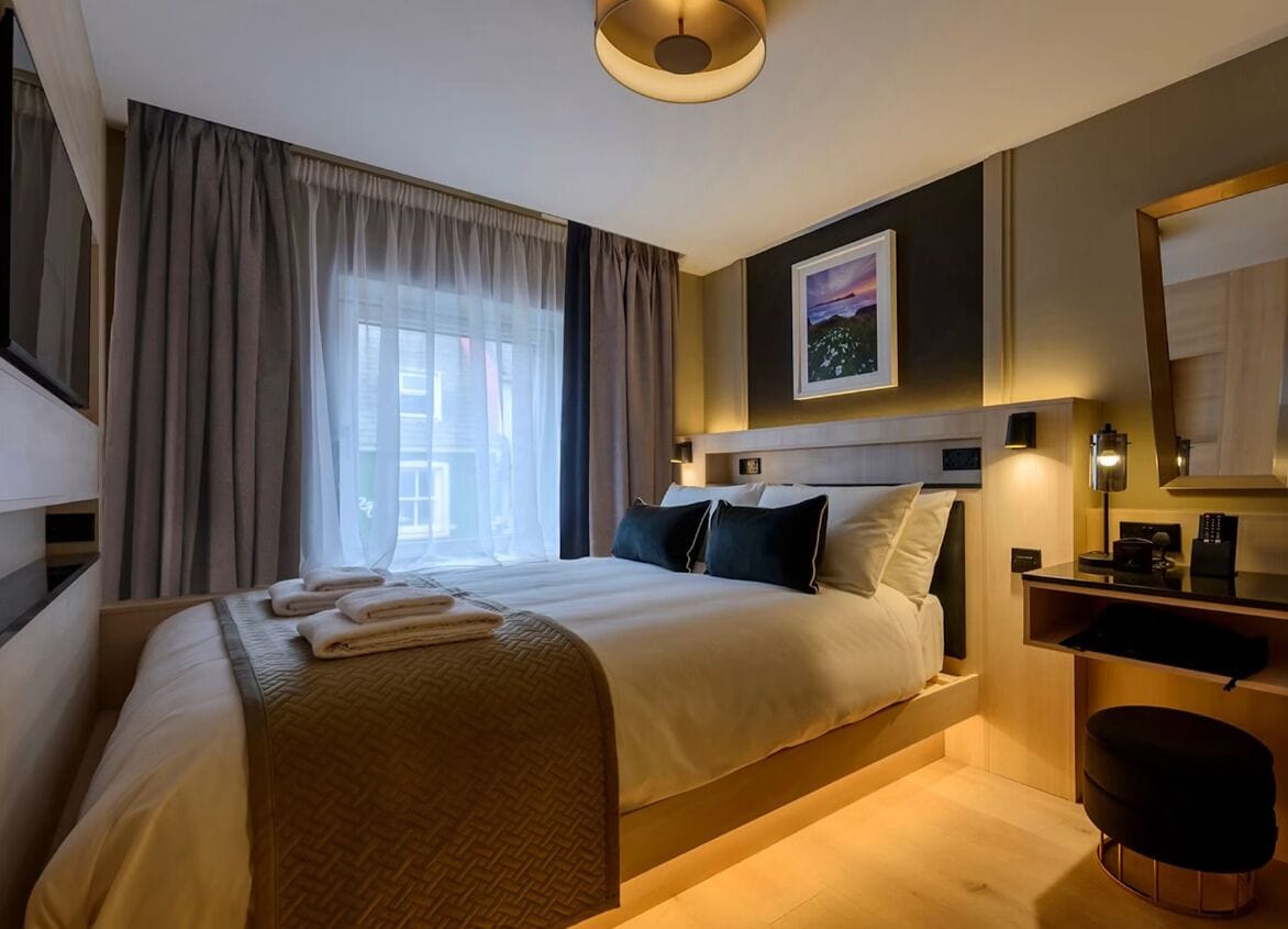 A hotel room with a bed, a desk, a chair, a large window with curtains, and a painting on the wall.