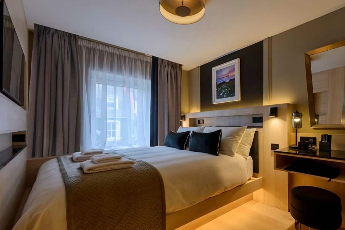 A hotel room with a bed, a desk, a chair, a large window with curtains, and a painting on the wall.