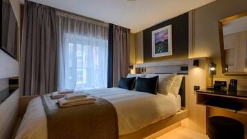 A hotel room with a bed, a desk, a chair, a large window with curtains, and a painting on the wall.