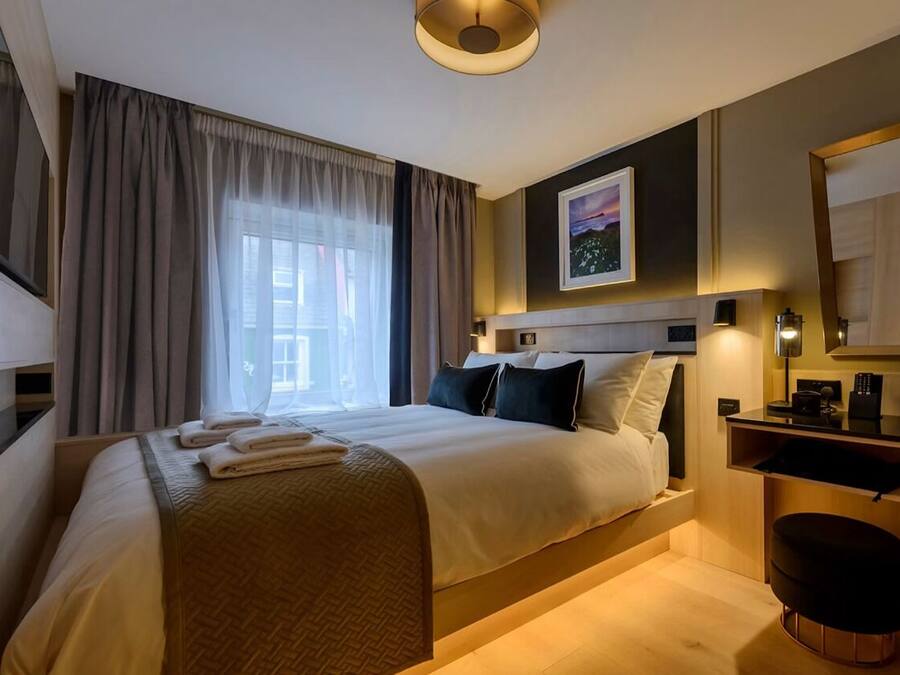 A hotel room with a bed, a desk, a chair, a large window with curtains, and a painting on the wall.