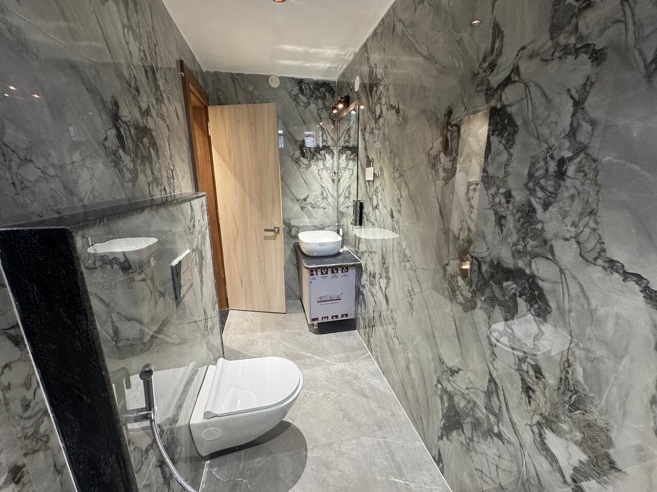 A modern bathroom with marble walls, a wooden door, a toilet, a sink, and a bidet.