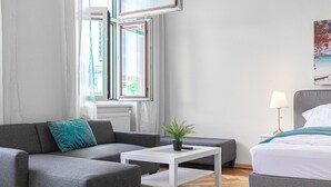 A modern hotel room with a grey sofa, a small white table with a plant, a grey armchair, a bed with white bedding, and a painting on the wall. - 2BR Apartment Steps From AKH Center (Vienna)