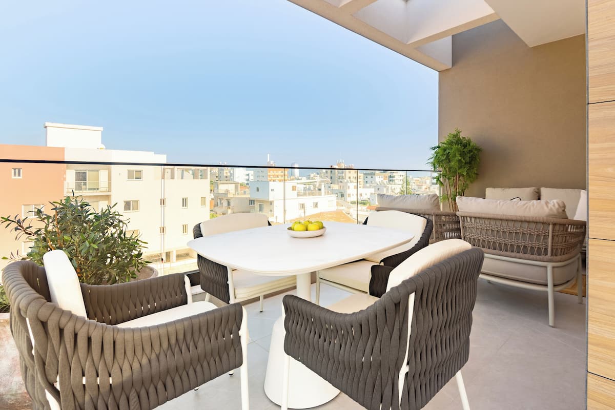 A balcony with a round table, four chairs, a potted plant, and a wicker sofa.
