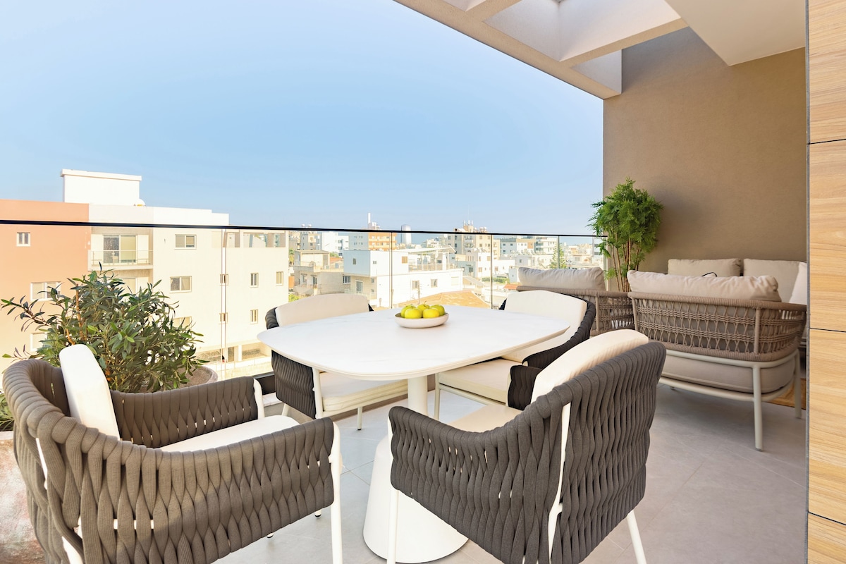 A balcony with a round table, four chairs, a potted plant, and a wicker sofa.
