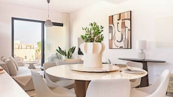 A modern living room with a round dining table, comfortable seating, a potted plant, and a large window offering a view.