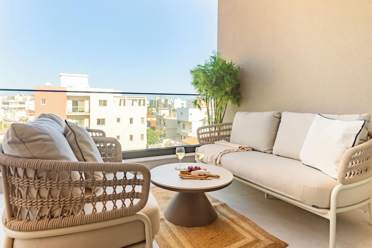 A balcony with a sofa, chairs, a table with a glass of wine, and a view of buildings.