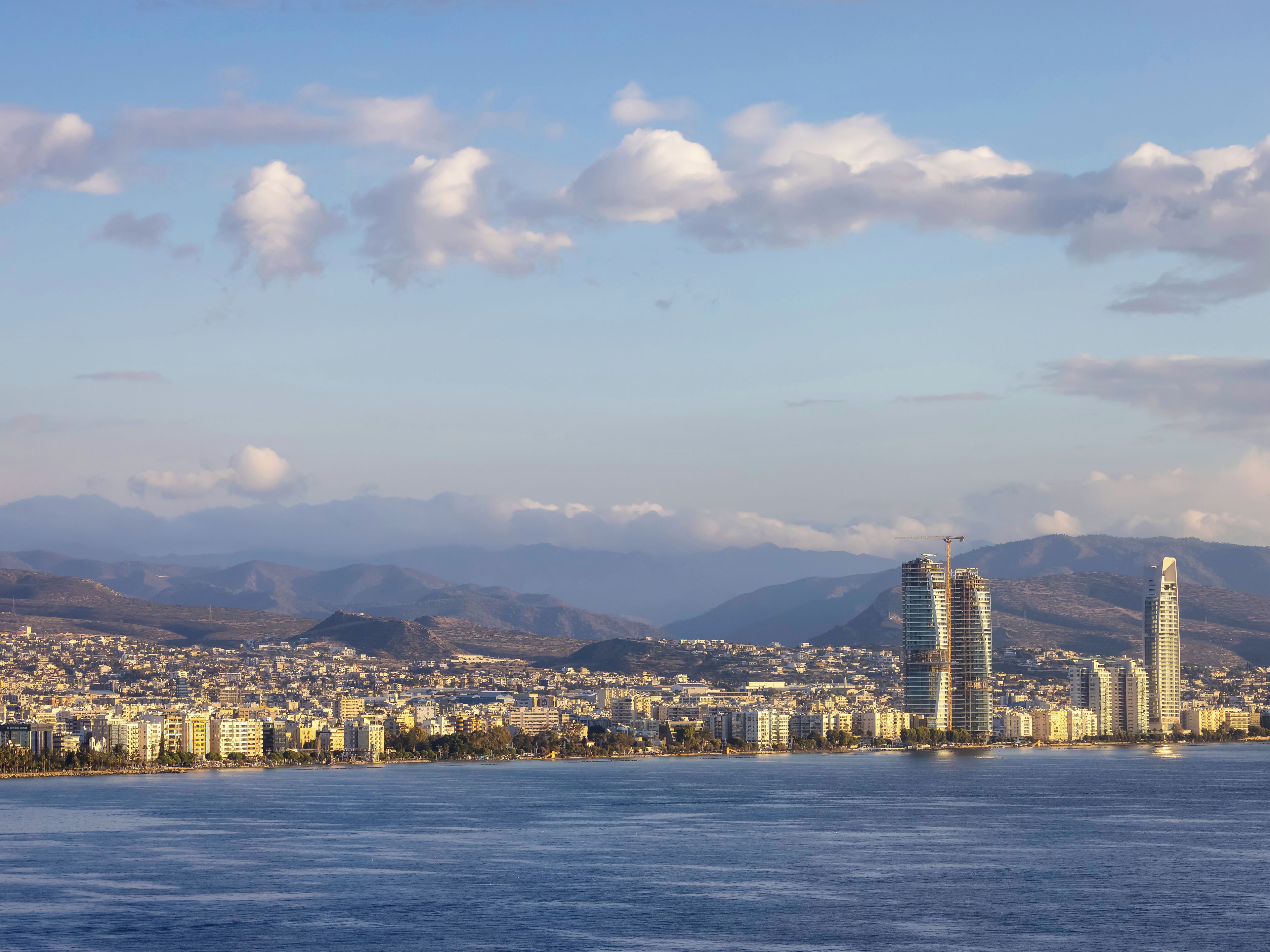 A coastal cityscape with modern high-rise buildings, a calm sea, and distant mountains under a partly cloudy sky.