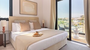 A hotel room with a large bed, a bedside table with a lamp, a view of the city through a large window, and a balcony with a chair. - Sanders City Boutique - Full Building (Limassol)