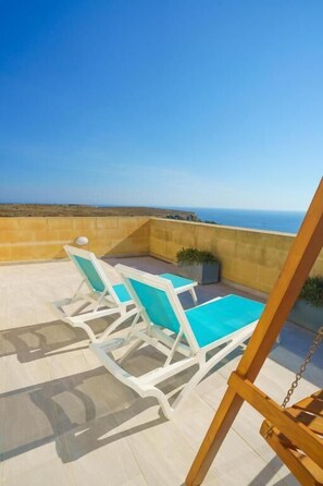 Eine Terrasse mit zwei weißen Liegestühlen mit türkisfarbenen Kissen, einer Holzschaukel und Blick aufs Meer.