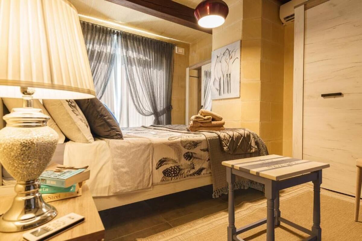 Une chambre à coucher avec un lit, une table de chevet avec des livres, une lampe, une petite table, un miroir et une fenêtre avec des rideaux.