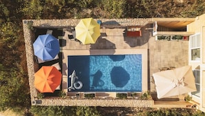 Vue aérienne d’un espace piscine avec plusieurs parasols, un plongeoir et un petit bateau.