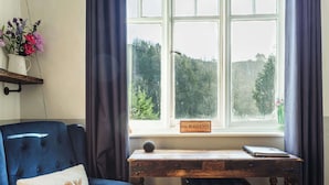 Une pièce chaleureuse avec un fauteuil bleu, une petite table en bois sur laquelle se trouve un ordinateur portable, et une fenêtre donnant sur un paysage verdoyant.