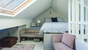 Une chambre mansardée chaleureuse, dotée d’un puits de lumière, d’un lit avec une literie à rayures, d’un banc, d’une chaise rose et d’une petite table avec une lampe.