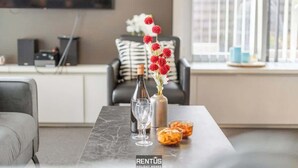 Ein modernes Wohnzimmer mit einem Marmortisch, einer Weinflasche und einer Vase mit roten Blumen. Ein Sofa mit gestreiften Kissen und ein an der Wand montierter Fernseher sind ebenfalls vorhanden.