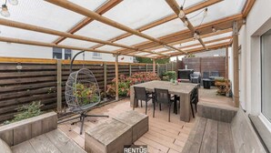 Ein überdachter Patio-Bereich mit einem Holztisch und Stühlen, einer hängenden Leuchte und einem Vogelkäfig.