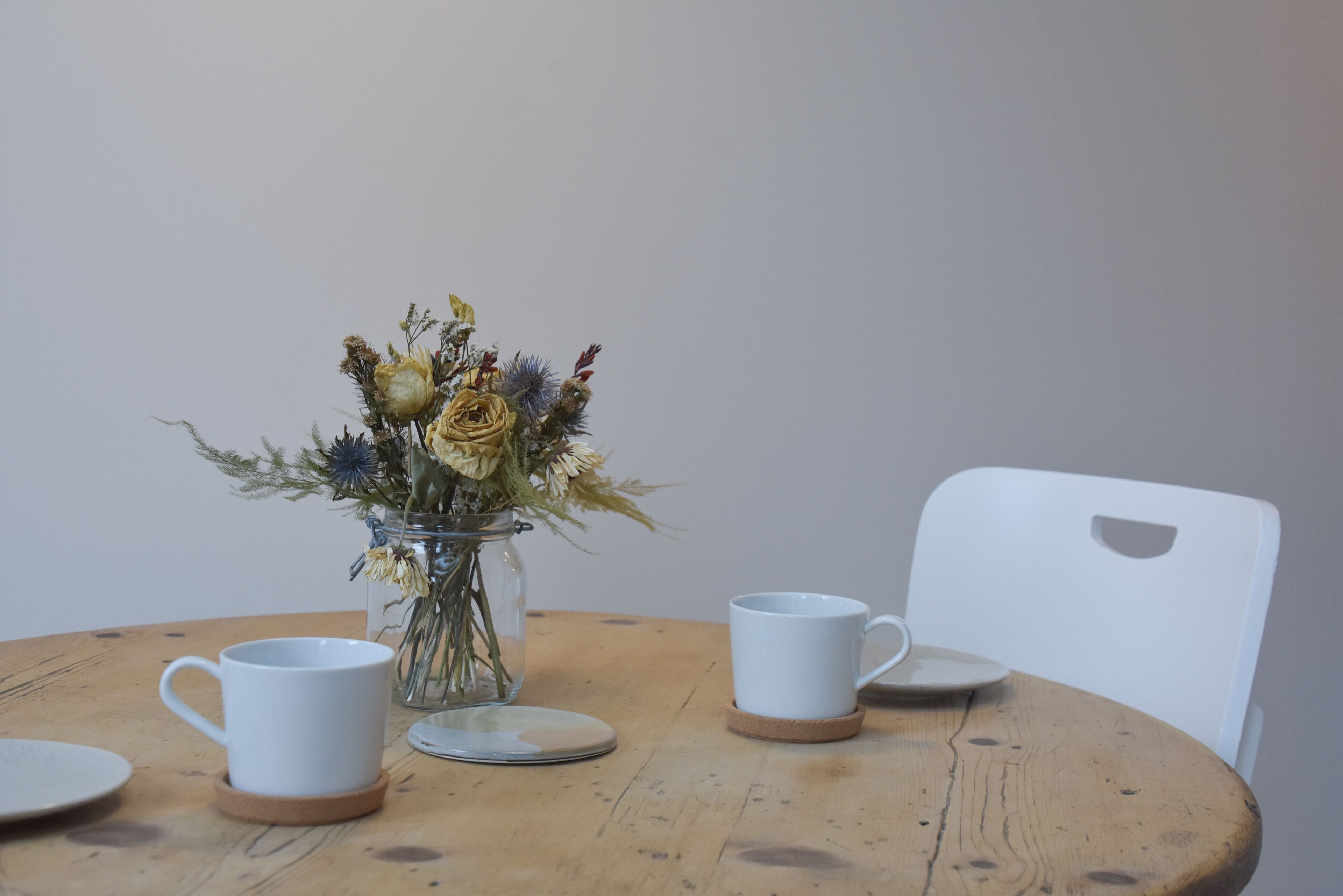 Een ronde houten tafel met een glazen vaas gevuld met gedroogde bloemen, twee witte keramische kopjes en een witte stoel.