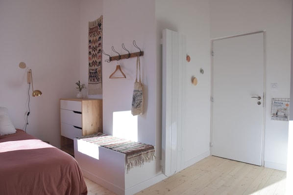 Une chambre avec un lit, une commode en bois, un banc avec une couverture, un porte-manteau avec un sac, une porte et un mur orné d’une affiche.