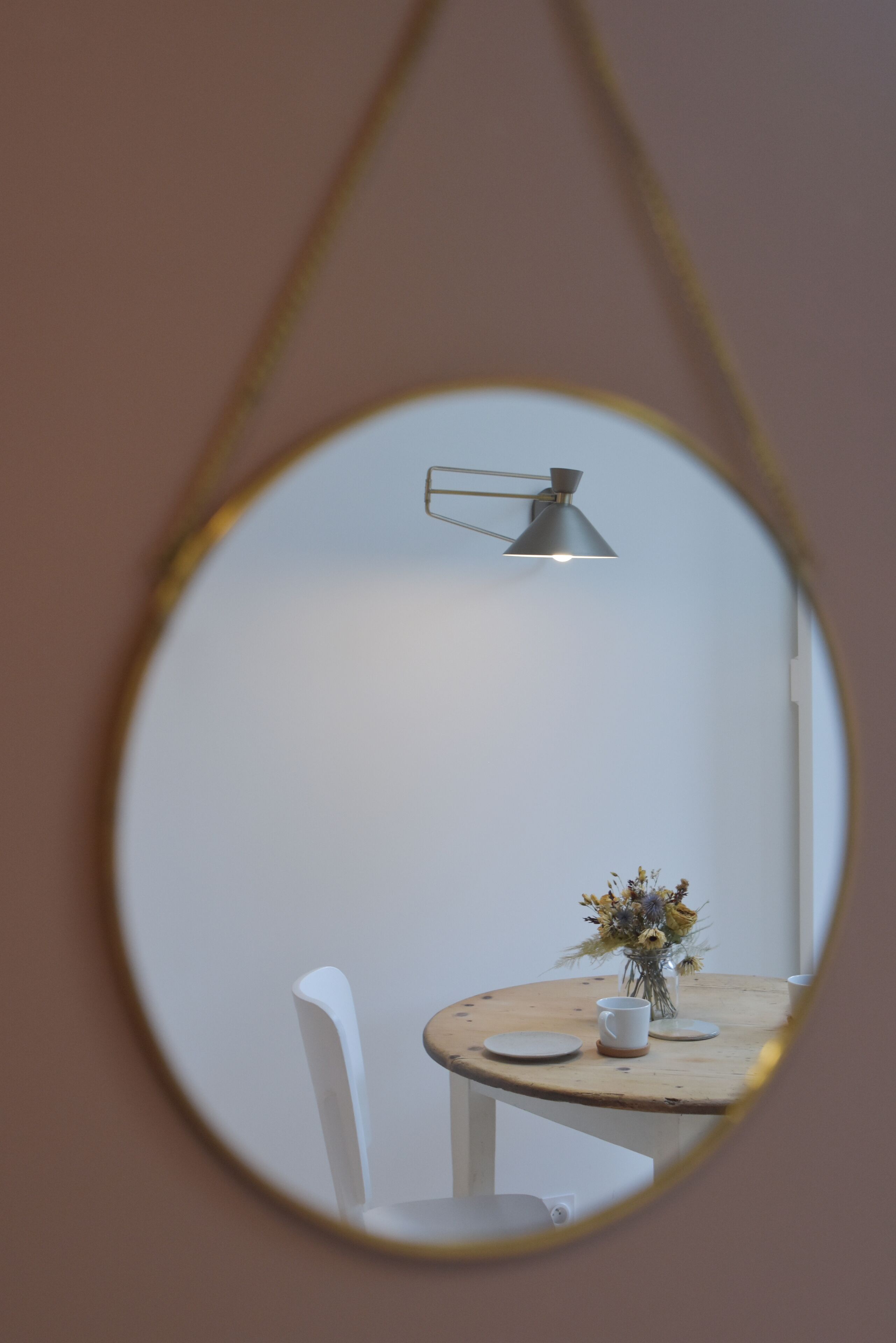 A round mirror reflecting a table with a vase of flowers, a white chair, and a hanging lamp.