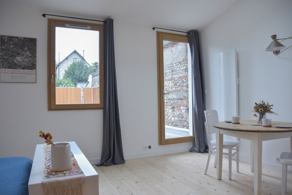 Une pièce avec un lit, une table sur laquelle se trouve un vase, et deux fenêtres avec des rideaux.