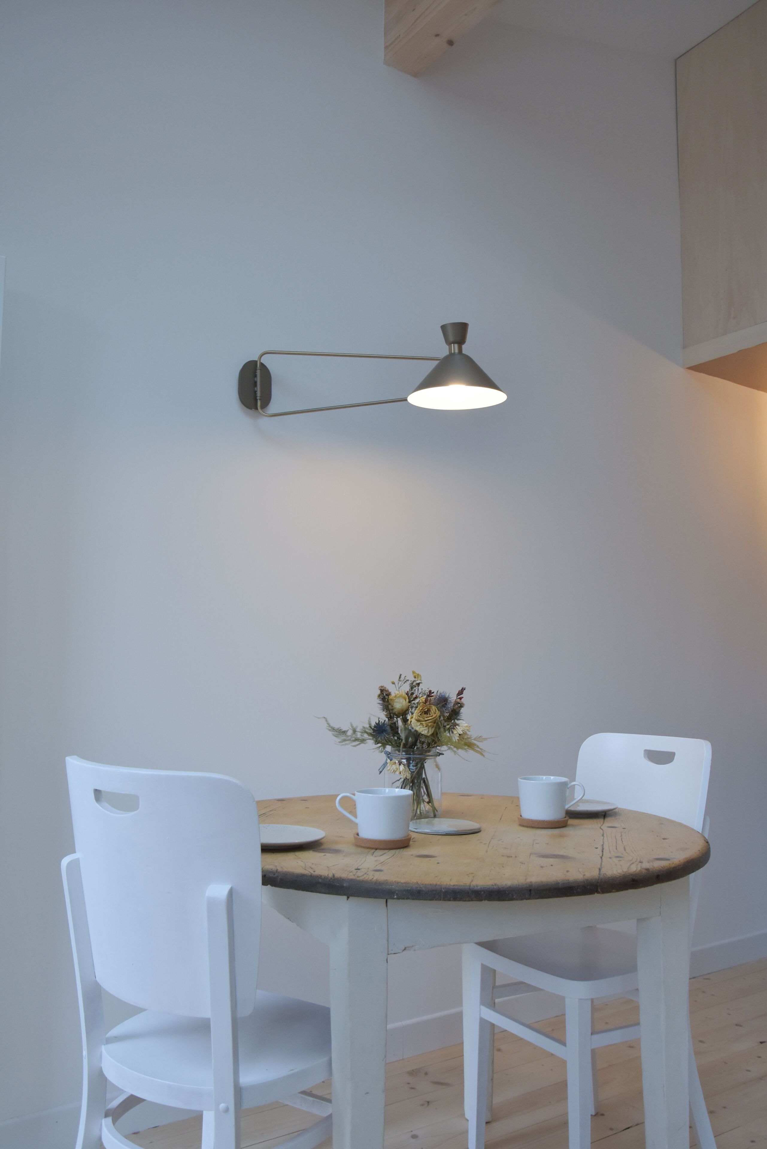 Comedor con mesa de madera, sillas blancas, un florero con flores y una lámpara fijada a la pared.