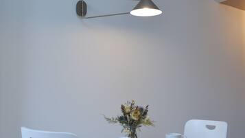 Comedor con mesa de madera, sillas blancas, un florero con flores y una lámpara fijada a la pared.