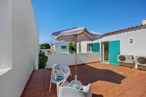 Une terrasse avec une chaise blanche, une petite table avec une bouteille et des verres, un parasol et des unités de climatisation.