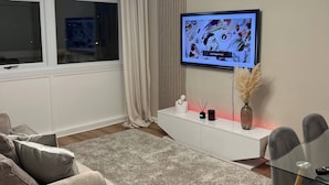 Un soggiorno moderno con un divano grigio, un tavolino da caffè in vetro, una TV a parete e un lampadario.