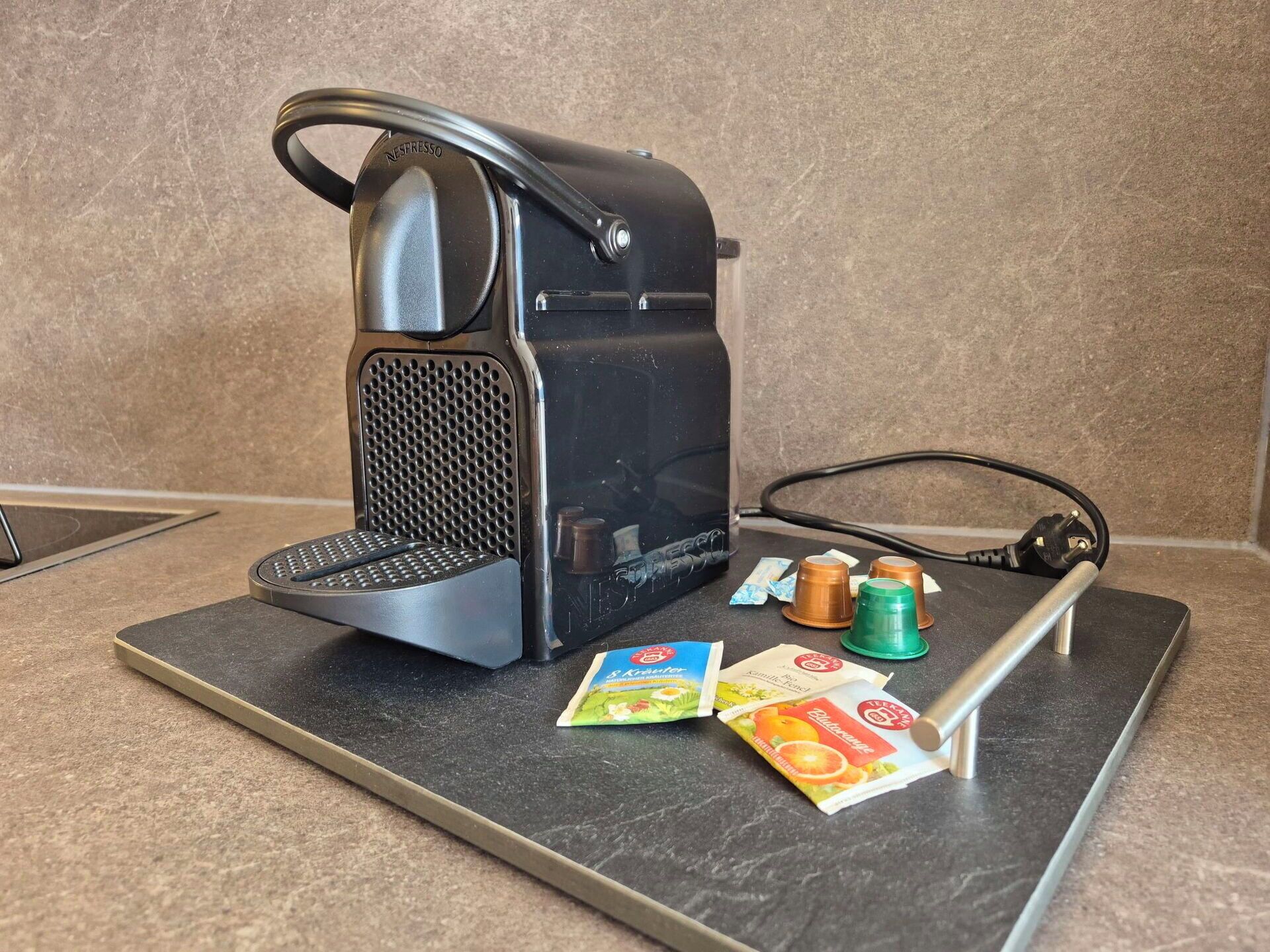 Nespresso-kahvinkeitin keittiön työtasolla, sen vieressä kahvikapselit ja metallinen teline.