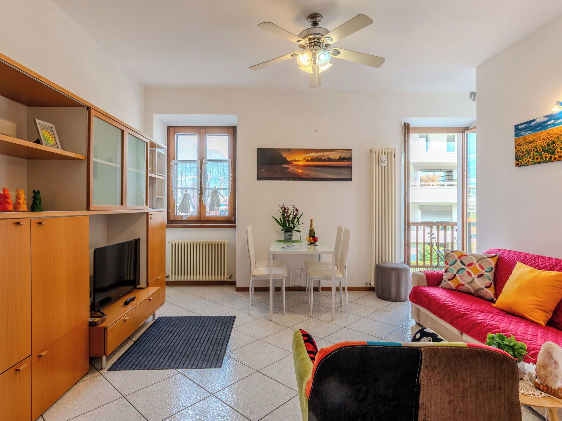 A living room with a TV, wooden cabinet, dining table, chairs, a red sofa, and a ceiling fan.