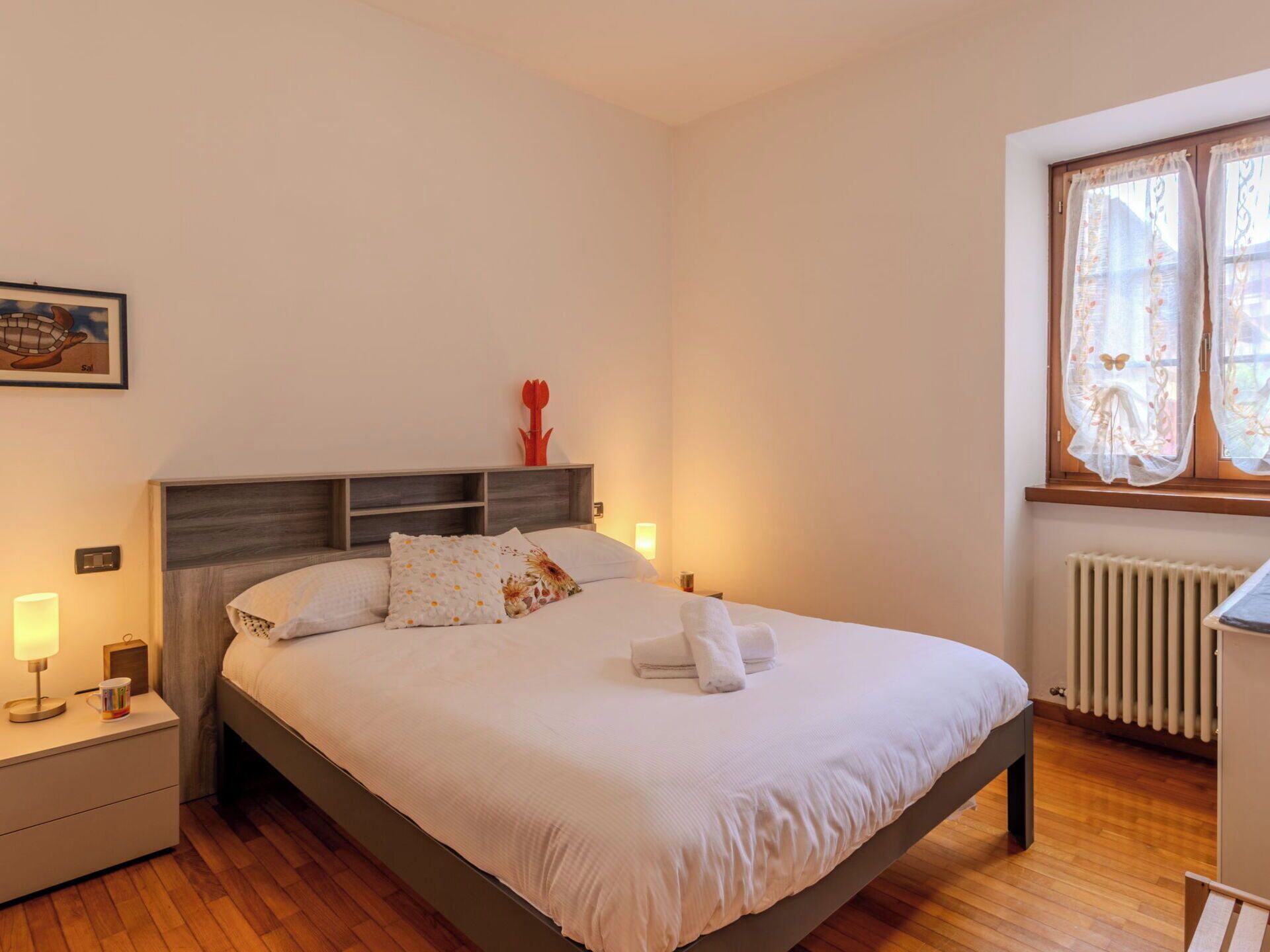 A bedroom with a bed, bedside lamps, a nightstand, a radiator, and a window with curtains.
