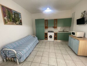 A compact kitchen with green cabinets, a washing machine, a microwave, and a bed with striped bedding. - Nice apartment for 7 guests with A/C, TV, balcony and pets allowed (Rosolina)