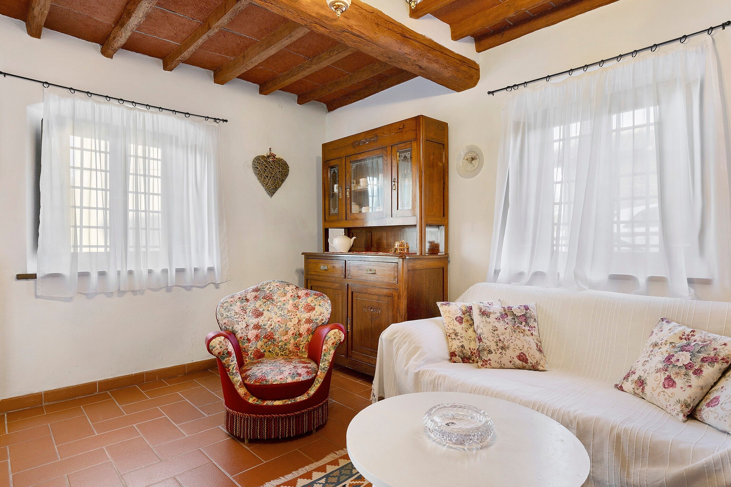 A room with a wooden ceiling, a floral armchair, a white sofa with floral cushions, a wooden cabinet, and a white table.