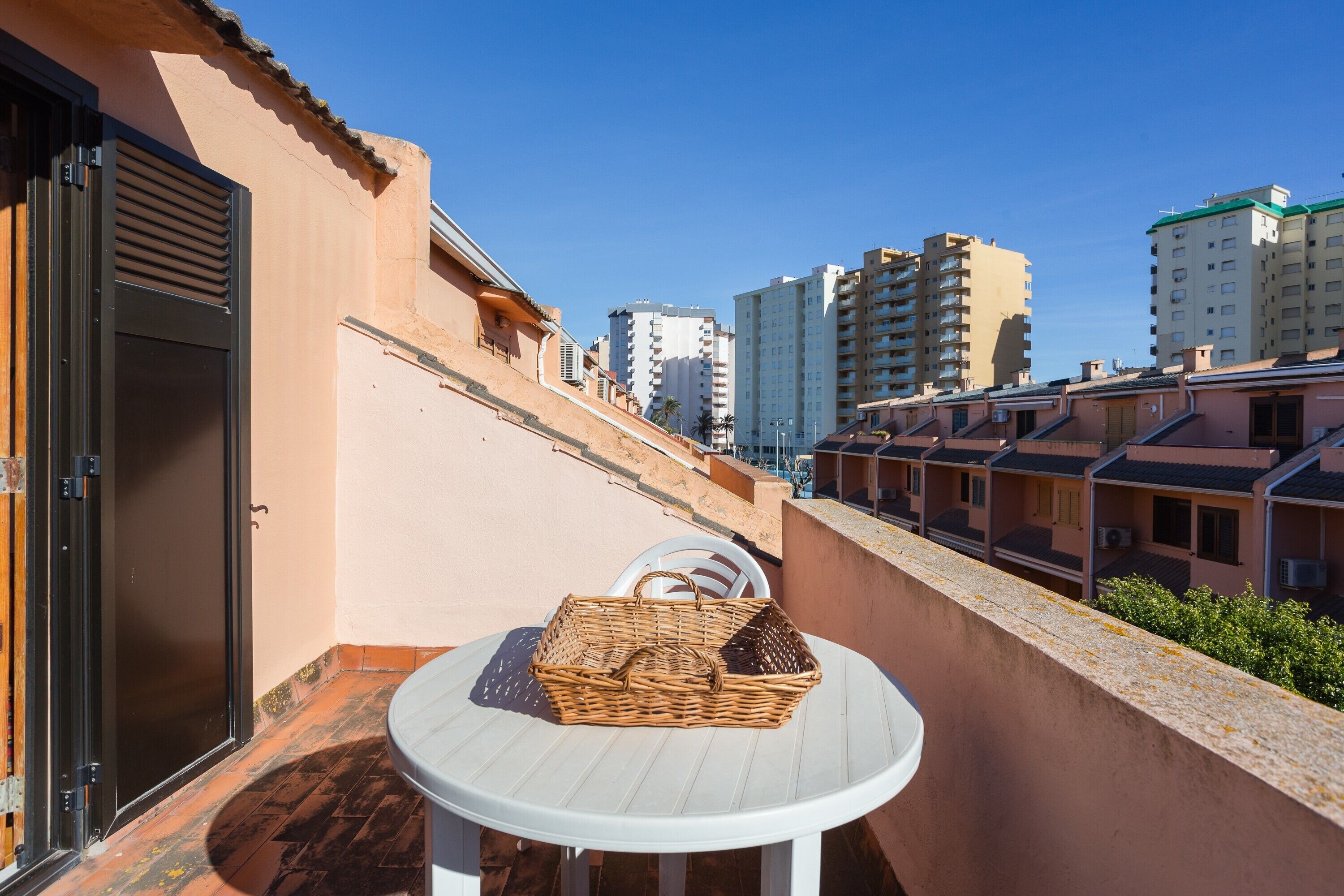 A balcony with a wicker basket on a white table, overlooking a row of buildings.