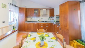Une cuisine avec des meubles de rangement en bois, une table à manger recouverte d’une nappe à motifs de tournesols et une fenêtre avec des rideaux.
