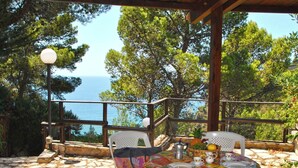 Uma área de estar coberta ao ar livre, com mesa posta para dois, com vista para o mar e árvores.