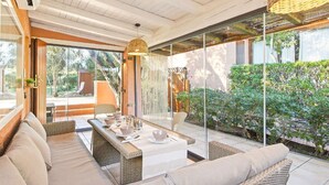 Un espace de vie extérieur moderne, doté d’un canapé, d’une table à manger et de chaises, entouré de portes vitrées et offrant une vue sur la verdure.