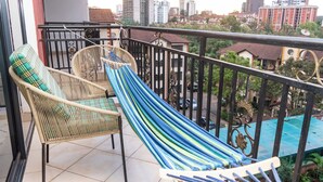 Ein Balkon mit Korbstühlen und einer Hängematte, mit Blick auf eine Stadtlandschaft mit Gebäuden und einem Swimmingpool.