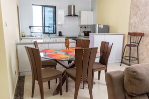 Una cocina moderna con gabinetes blancos, una campana extractora de acero inoxidable y una zona de comedor con mesa de vidrio y sillas de madera.