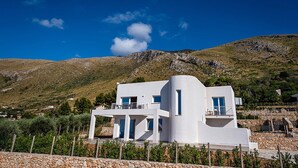 Een moderne witte villa met een gebogen gevel, een balkon en uitzicht op een bergachtig landschap.