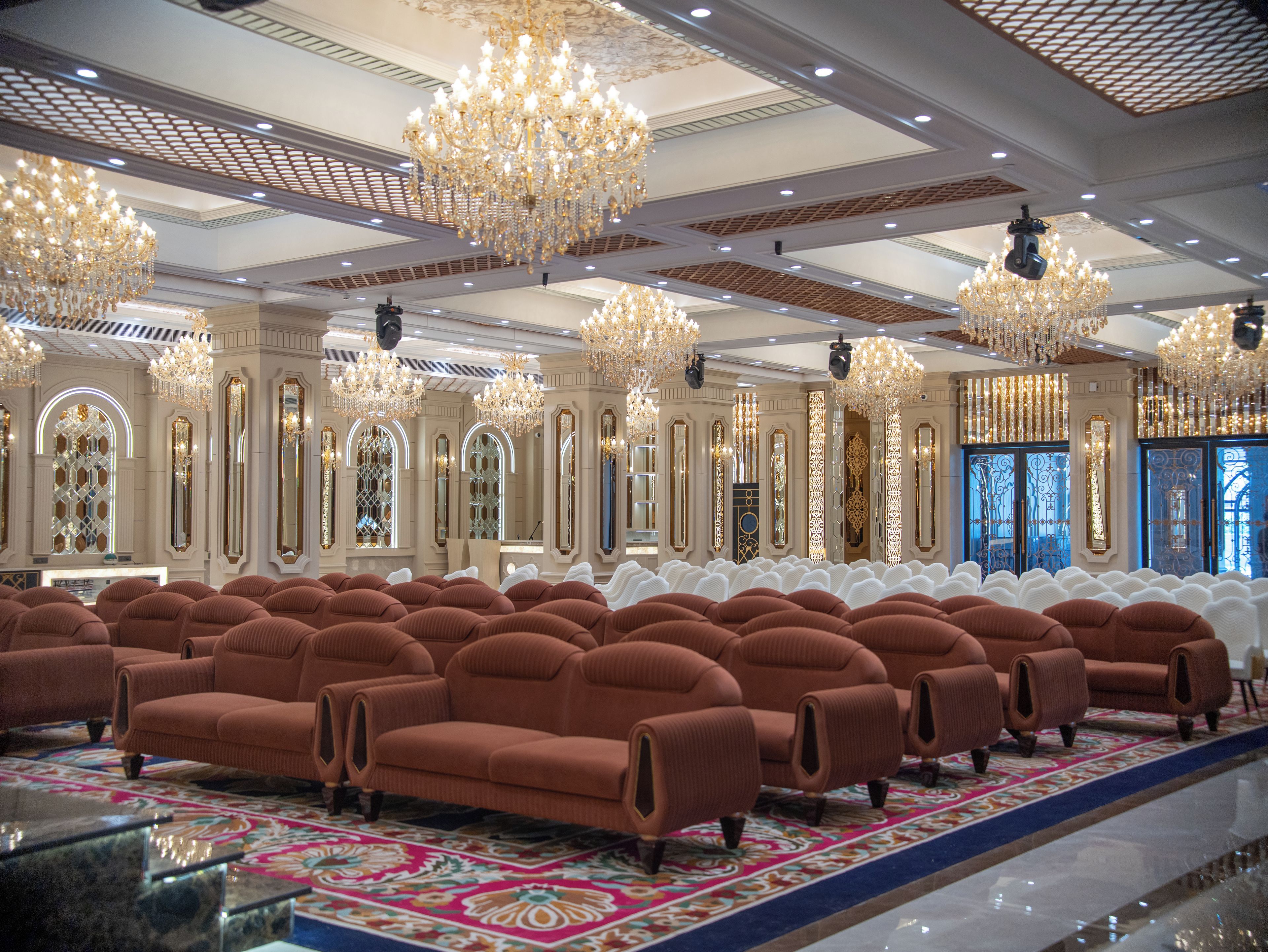 Un hallo espacioso con filas de sofás marrones, candelabros ornamentados y una alfombra estampada.