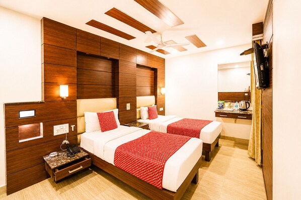 A hotel room with two beds, wooden headboards, a flat-screen TV, a bedside table with a lamp, and a small kitchenette area. - Hotel Quadis - East Of Kailash (New Delhi)