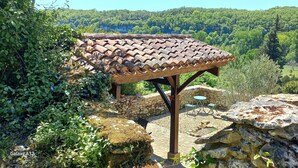 A stone pergola with a tiled roof in a garden setting, surrounded by greenery and a mountainous backdrop. - LITTLE HAVEN OF PEACE (Les Eyzies)