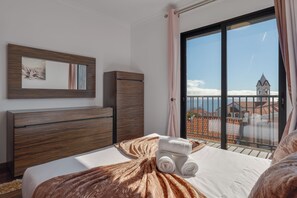 A hotel room with a bed, a wooden dresser, a mirror, and a balcony with a view of a church and buildings. - Andrade's Villa by HRM (Santa Cruz)