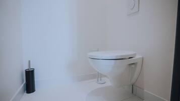A white toilet in a bathroom with a black toilet brush and a wall-mounted flush handle.