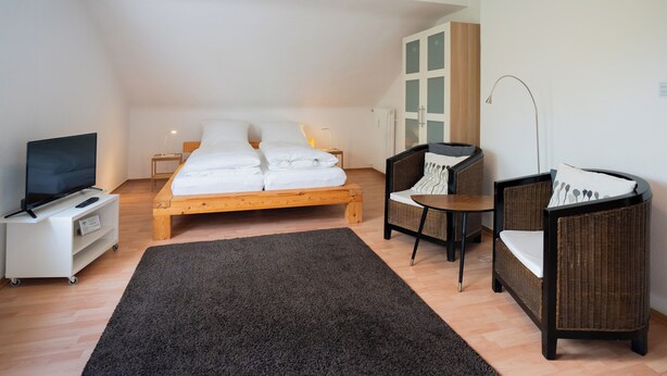 A bedroom with a wooden bed, a TV on a white stand, a small table, and a wicker chair. - Guesthouse Elena Apartment 21 (Kassel)